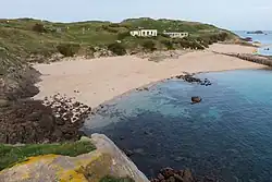 Photographie en couleur montrant une plage de sable, avec à gauche une pente recouverte de végétation et de roches. Un petit bâtiment carré et un autre plus grand sont visibles au centre. Au fond, on devine un grillage et des panneaux signalant l'interdiction d'aller plus loin.