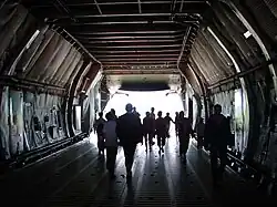 Plusieurs personnes en contre-jour debout dans la soute, la rampe arrière de l'avion est ouverte.