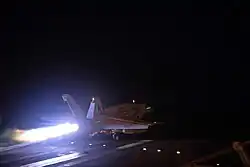 Photo de nuit d'un avion de chasse catapulté depuis un porte-avion. La scène est éclairée par la post-combustion des réacteurs, le reste du pont d'envol est plongé dans le noir.