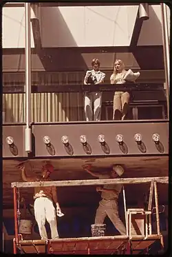 Photographie des années 1970 montrant deux ouvriers travaillant en sous-face de passerelle tandis que deux personnes se tiennent à l'intérieur de la passerelle vitrée.