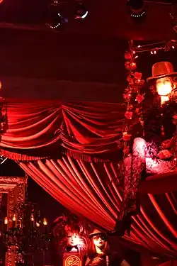 Photo en couleur d'un lieu sombre avec des points de lumière et où se distinguent des draperies rouges, des guirlandes, quelques silhouettes