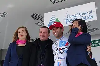 De trois-quarts, quatre personnes sourient. Un coureur au maillot blanc serre les personnes à côté de lui.