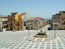 Photographie couleur d’une place de village avec une fontaine au centre.
