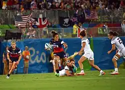 Au centre, de face, une athlète avec le ballon sous le bras droit. Avec le bras gauche, elle s'apprête à repousser une adversaire.