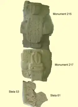 Plusieurs fragments de sculptures cassées alignées comme un pilier vertical, avec un personnage humain près de la tête d'une chauve-souris géante, elle-même reposant sur une sculpture très endommagée montrant des motifs complexes taillés en relief.