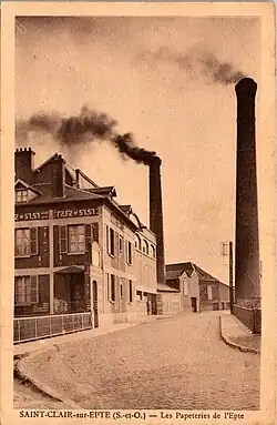 Une rue d'usine avec, au second plan, deux cheminées fumant. Carte postale sépia.