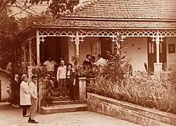 Photographie en noir et blanc où des touristes visitent une maison. Il n'y a que la partie gauche du bâtiment qui est visible. Sept personnes se trouvent sous le porche, deux autres sont debout sur un petit escalier à l'entée du porche. Deux personnes sont à côté de l'escalier.