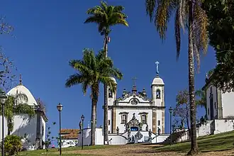 Photographie en couleurs. Vue lointaine de l'église au centre, avec une chapelle à gauche et à droite et des palmiers au premier plan.