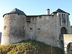 Vue de l’accès d’un château fort, avec une tour ronde à gauche et une tour carrée surplombant l’entrée avec pont-levis.