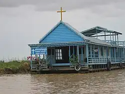 Le bâtiment est de couleur bleue avec une croix au sommet du toit au-dessus de l'entrée. À gauche, un panneau avec l'inscription église catholique de Chong Khneas en khmer et en anglais.
