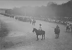 Photographie en noir et blanc de cavaliers au premier plan devant des troupes militaires.