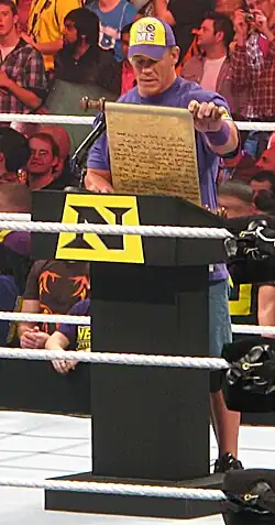 John Cena, nouveau membre du Nexus, se tient debout sur le ring, en face d'un pupitre. Il lit un discours rédigé sur un parchemin.