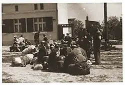 Une vingtaine d’hommes sont assis au sol ou sur leurs bagages, entourés de paquets, en extérieur. Plusieurs policiers, avec des brassards et des képis, les entourent, debout. En arrière-plan, quelques immeubles.
