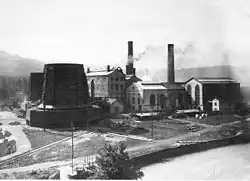 Photo noir et blanc montrant de hauts bâtiments industriels accompagnés de deux tours de refroidissement métalliques et de deux cheminées d'usine.