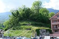 Petite colline peuplée d'arbustes et des hautes herbes, avec une construction en pierres grises et en ciment à sa base.