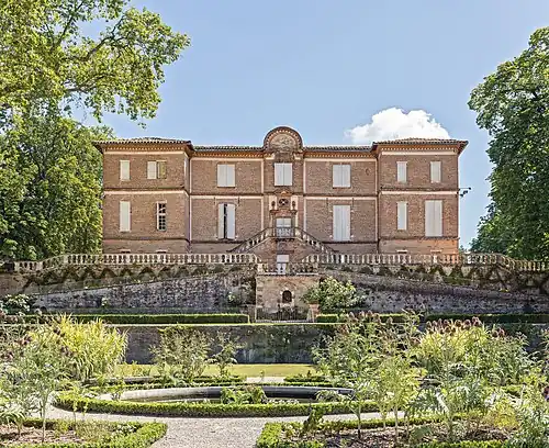 La photo couleur présente des terrasses fleuries au premier–plan et un château style classique en briques rouges.