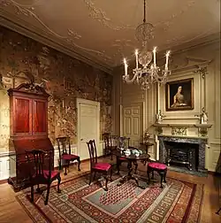 Intérieur d'une pièce de style ancien, probablement américaine du dix-huitième siècle, avec des meubles en bois brun rouge autour d'une table ronde. Des chaises et tabourets rouges contrastent avec un tapis aux motifs complexes. Les murs sont recouverts de papier peint oriental, et un lustre en cristal ajoute du luxe. Une peinture d'une femme est accrochée au-dessus de la cheminée.