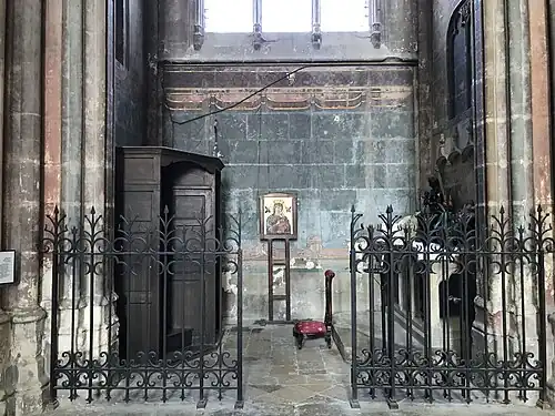 Pièce aux murs partiellement peints et accessible par une grille en fer forgé ouverte. À l'intérieur, un confessionnal, une icône accrochée au mur, un prie-Dieu et un autel.