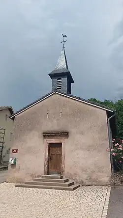 Chapelle Saint-Bernard à Flatten vue de face. Elle possède une porte en bois accessible par quatres marches d'escalier. Ses murs sont beiges et son toit est fait de tuiles grises. Un petit clocher surmonte la porte, sur son toit en pointe se trouve une croix en fer elle-même surmontée d'une girouette en forme de coq.