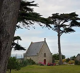 Vue d’une chapelle aux murs de granit et toit en ardoises, avec un arbre en premier plan