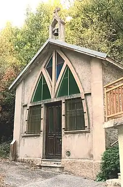 Photographie d'une chapelle en lisière de forêt, avec des vitraux verts et bleus ainsi que la porte marron en bois.
