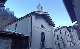 Photographie d'une chapelle entourée de maisons avec son clocher rénové et sa porte grillée.