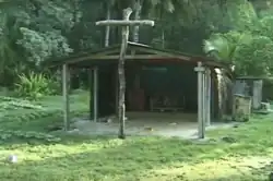 Photo d'un bâtiment en bois, sans mur sur le devant avec à l'entrée une grande croix en bois, à l'arrière-plan la forêt.