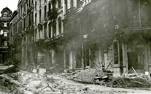 Photo ancienne en noir et blanc d'une rue avec des bâtiments ruinés. Des débris jonchent la chaussée.