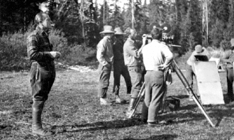 Photographie en noir et blanc d'un groupe d'hommes rassemblés devant une caméra sur pied, en extérieur ; personnage principal à l'écart.