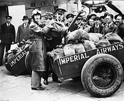 Photographie noire et blanc de Kingsford Smith tenant un sac de courrier, des chariots remplis d'autres sacs derrière.