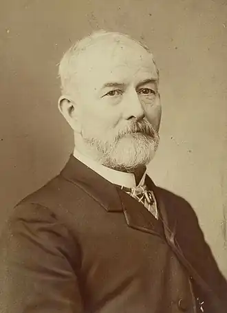 Photographie en noir et blanc avec une teinte marron. Elle représente un homme blanc, qui porte une barbe blanche et une veste de costume foncée.