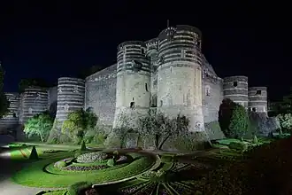  Photographie d'un château éclairé la nuit, six tours imposantes et des jardins au premier-plan dans les douves.