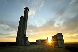 photo à contre-jour des ruines d'une construction en pierre. À gauche, une tour surmontée d'une tourelle carrée a gardé son aspect. À droite, les murs sont tronqués. Le soleil levant s'immisce dans une trouée des remparts.