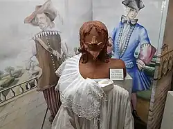 Photographie d'une vitrine. À l'arrière-plan, un fond illustré montre un homme en costume beige de mousquetaire du XVIIe&nbsp;siècle avec bandoulière, fraise en dentelles et chapeau à plume, et un autre en costume du XVIIIe&nbsp;siècle, bleu à manches violettes, portant une perruque blanche et un tricorne noir. Au premier plan, un mannequin en bois sculpté représentant un homme à moustache et barbiche fine à la façon du XVIIe&nbsp;siècle expose une large chemise blanche au col orné de dentelles.