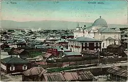 Ancienne carte postale en couleur. Au premier plan, se distingue des maisons en bois aux toits pentus, serrées les unes contre les autres. Au centre, un grand bâtiment religieux se dresse avec un dôme proéminent qui domine le paysage. En arrière-plan, la ville s'étend à perte de vue, avec des collines et ses nombreux bâtiments. En haut à gauche est écrit Tchita en russe.