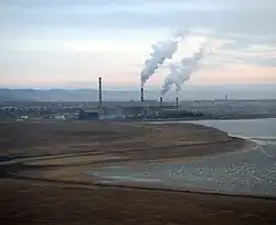 Photographie depuis un avion volant à basse altitude d'un lac avec derrière quatre cheminées (deux grandes, deux petites), avec trois qui rejettent de la fumée. Le paysage est pris à l'aube, avec un ciel légèrement coloré en arrière-plan.