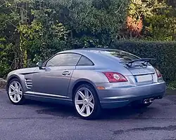 Une voiture grise, 2 portes, garée devant une haie et vue de l'arrière gauche.