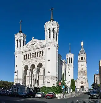 Photographie couleur montrant la basilique, au sommet de la colline, avec ses quatre tours et la statue de la Vierge, prise depuis le centre-ville de Lyon.
