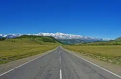 Photographie depuis le centre de la route. Le paysage est verdoyant, avec en arrière-plan des cimes enneigées, dans la steppe de Kouraï.