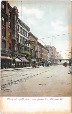 photo colorée d'une large avenus bordée d'immeubles avec beaucoup de boutiques, la foule sur les trottoirs, et deux voitues à cheval.