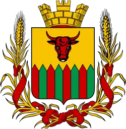 Bblason, comprenant un écu jaune avec une tête de taureau rouge en son centre, surmonté d'une couronne murale dorée. L'écu est orné en bas de motifs alternés rouges et verts, évoquant une forme géométrique en zigzag. Le tout est entouré de branches de blé dorées, nouées par des rubans rouges, ajoutant un symbole de prospérité et de tradition agricole.