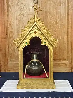 Photographie en couleurs d'une fiole en verre décorée d'un ruban dans un reliquaire en cuivre vitré.