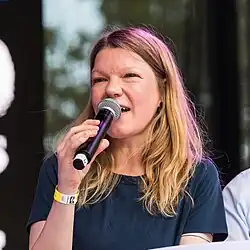 photo d'une femme en shirt noir et qui parle dans un micro tenu par sa main droite.