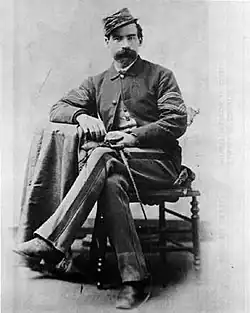 Portrait d'un homme en habits militaires, assis, portant la moustache et coiffé d'une casquette militaire.