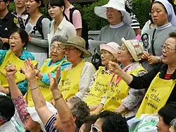 Anciennes femmes de réconfort, aujourd'hui très âgées, alignées et assises lors d'une manifestation, elles arborent des tabliers sur lesquels sont écrites des revendications.