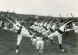 Photographie montrant des gymnastes en exercice, soulevant une barre entre les bras, une jambe relevée.