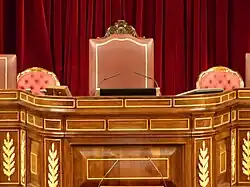 Photo d'une tribune en bois surmontée de trois fauteuils de couleur rose, à l'arrière-plan, un rideau rouge