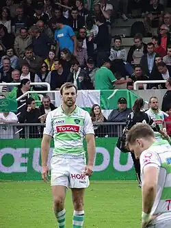 Photo en couleur montrant un joueur de rugby habillé en blanc et vert marchant sur une pelouse, au fond des spectateurs.