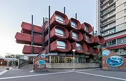 Une photographie du conservatoire de Montreuil prise en contre-plongée. Le bâtiment se distingue par son architecture originale, évocatrice du style des années 70 : des sortes de "cellules" tubulaires, de couleur rouille et dotées de vitres à leurs extrémités, constituent le bâtiment sur 3 étages.