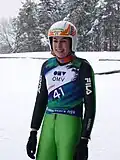portrait jusqu'à mi-cuisses de Evelyn Insam souriante, en combinaison de saut à ski verte, coiffée de son casque, avec un dossard de compétition numéro 41, sur fond de paysage hivernal avec des sapins